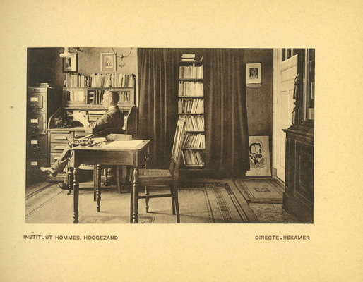De directeurskamer met de heer Hommes zittend aan een bureau. Op de achtergrond verschillende boekenkasten en op de voorgrond een lessenaar met stoel.