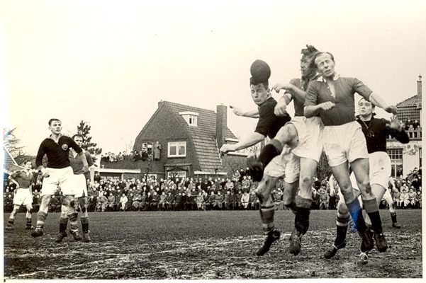 Een spelmoment uit een wedstrijd in 1952 met een drietal mannen die naar de bal springen en veel publiek.
