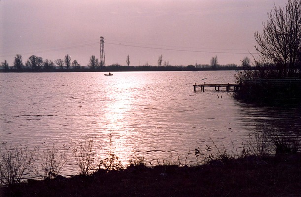Foxholstermeer gezien vanaf de Scheepswervenweg. Op het water een klein bootje en de schitterende zon. Rechts een klein steigertje. Op de achtergrond de spoorbaan.