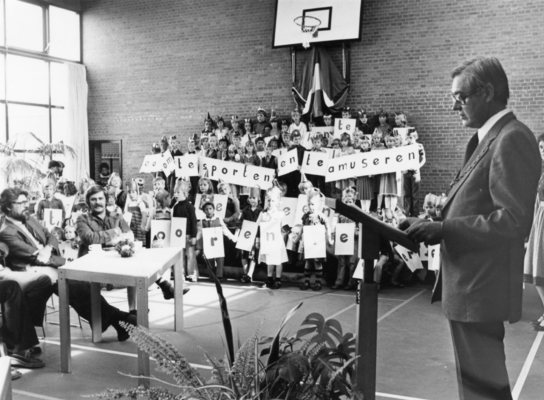 De opening van de school in de gymzaal. De burgemeester houdt een toespraak en op de achtergrond staan kinderen te poseren.