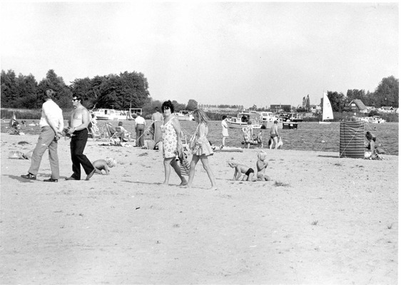 Het strand bij Meerwijck in de jaren zeventig, met langslopende mensen, spelende kinderen en mensen die in de zon liggen. Op de achtergrond liggen verschillende bootjes in het water.