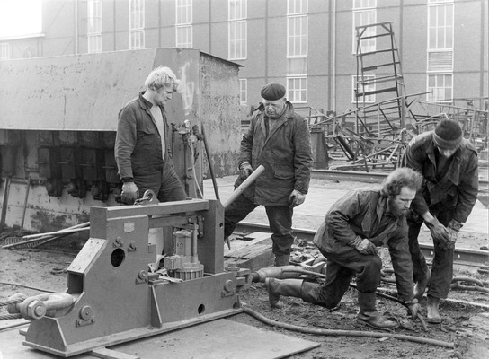 Foto genomen bij de scheepswerf E.J. Smit. Op de voorgrond vier mannen die aan het werk zijn. Waarmee is niet precies duidelijk. Op de achtergrond allerlei ijzerwerk en het hoge gebouw van de werf.